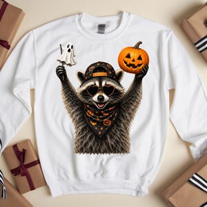 May include: A white long-sleeve sweatshirt featuring a graphic of a raccoon wearing a black baseball cap, heart-shaped sunglasses, and a black bandana with orange pumpkins. The raccoon holds a small white ghost on a stick and an orange jack-o'-lantern.