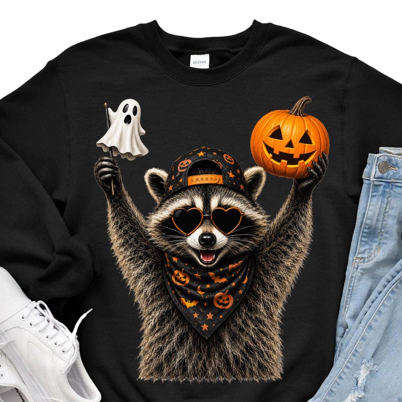 May include: A black sweatshirt featuring a graphic of a raccoon wearing a black baseball cap with orange pumpkins, heart-shaped sunglasses, and a black bandana with orange pumpkins and bats. The raccoon holds a white ghost and an orange jack-o'-lantern.