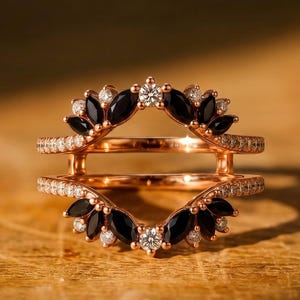 May include: A rose gold ring guard featuring two curved bands connected by a bridge. The design is adorned with marquise-cut black stones and small round white stones arranged in a fan-like pattern along the top and bottom edges of the ring.