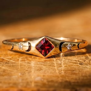 Peut inclure: Une bague en or ornée d'une pierre précieuse rouge foncé taille princesse au centre. L'anneau est décoré de croissants de lune gravés et de deux petites pierres rondes claires serties en forme d'étoile de chaque côté de la pierre rouge.