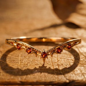 May include: A gold V-shaped ring featuring a central star-set red gemstone flanked by four smaller red stones. The band incorporates crescent moon motifs and delicate engraved detailing along the metal surface, creating a pointed chevron silhouette.
