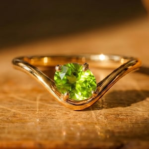 May include: A gold ring featuring a single, round, faceted, bright lime-green peridot gemstone set in a V-shaped band. The metal band has a polished finish, and the gemstone is secured by four small prongs, catching the light to show its clarity.