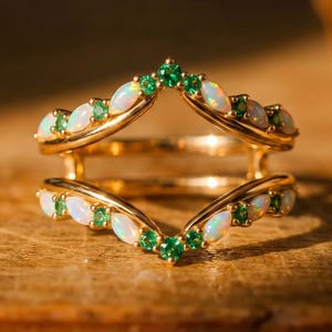 May include: A gold ring guard featuring two chevron-shaped bands connected at the sides. The bands are set with alternating marquise-cut opals and round-cut green gemstones, creating a symmetrical V-shaped design with a polished metallic finish.