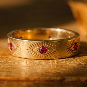 Könnte beinhalten: Ein goldfarbener Ring mit einem eingravierten Augendesign und roten Edelsteinen. Der Ring hat mehrere Augenmotive mit einem zentralen roten Edelstein in jedem Auge. Der Ring liegt auf einer Holzoberfläche.