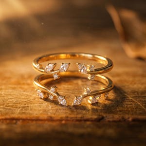 May include: Two gold rings stacked on top of each other, each adorned with small, clear gemstones in a leaf-like pattern. The rings are set against a warm, wooden background, creating a luxurious feel. The gemstones sparkle in the light.