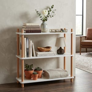 May include: A three-tier shelving unit featuring white rectangular shelves supported by four light-toned wooden cylindrical posts. The shelves hold various items including stacked books, a ceramic vase with flowers, a small lamp, potted succulents, and a folded textile.