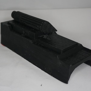 May include: A black, rectangular object with a cylindrical top and a textured surface. The object has a stepped design and a curved base. The overall shape suggests a mechanical or industrial component.