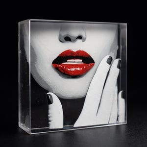 May include: A clear acrylic block featuring a black and white image of a face with bright red lips and a hand with black nail polish. The text "Corazón de Jade" is at the bottom.