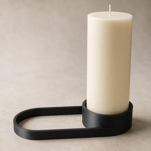 Puede incluir: Una vela de pilar crema se encuentra en un portavelas negro de forma ovalada. La vela tiene una mecha visible. El portavelas tiene un diseño moderno y minimalista.