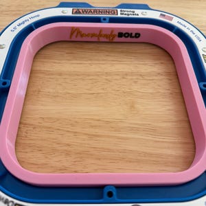 Op de afbeelding: Een blauwe en roze borduurring met de tekst "Marselly BOLD". De ring is gemarkeerd met "5.5 Mighty Hoop" en "Made in the USA". De ring heeft een waarschuwingslabel voor sterke magneten.