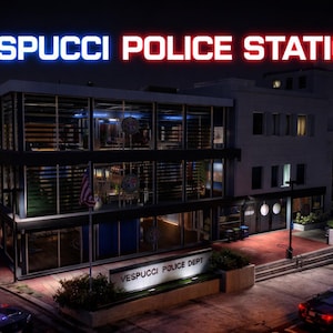 May include: Nighttime exterior shot of the Vespucchi Police Station. The building is a modern two-story structure with large glass windows. The words "VESPUCCI POLICE STATION" are illuminated in neon lights. Police cars with flashing lights are parked in front.