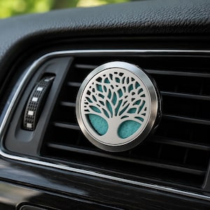 Peut inclure: Diffuseur d'air pour voiture argenté, orné d'un motif d'arbre de vie. Le diffuseur circulaire révèle un tampon turquoise à travers le motif. Fixé à une grille d'aération noire, il parfume l'intérieur du véhicule.