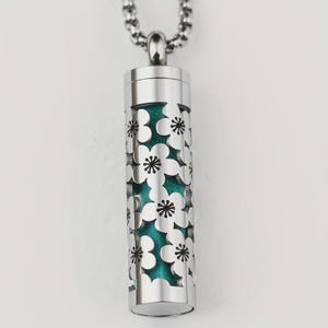 Peut inclure: Un collier pendentif argenté avec un charm cylindrique. Le charm présente un motif floral découpé révélant un intérieur turquoise. La chaîne du collier est également argentée. Le pendentif est probablement en acier inoxydable.