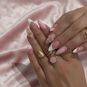 Puede incluir: Primer plano de uñas largas en forma de almendra con un diseño rosa y blanco. Algunas uñas presentan adornos florales en 3D y detalles dorados. Las manos también muestran anillos dorados, uno con una piedra rosa y otro con un emblema religioso.