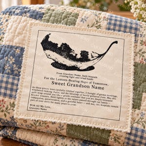Puede incluir: Una colcha de retazos con cuadrados florales y de cuadros vichy en azul, verde y crema. Una etiqueta rectangular de tela está cosida en el centro, mostrando una ilustración de una hoja negra y el texto: From Grandma Name, For the Lantern-Bearing Hope of Tomorrow, Sweet Grandson Name, With All My Love, Grandma Name, y un mensaje largo.