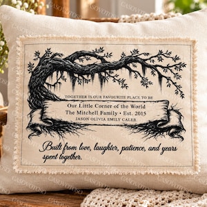 Op de afbeelding: Een vierkant beige sierkussen met een stoffen patch met gerafelde randen en een zwarte inktillustratie van een uitgestrekte boom. De tekst luidt: Together is our favourite place to be, Our Little Corner of the World, The Mitchell Family, Est. 2015, Jason Olivia Emily Caleb, Built from love, laughter, patience, and years spent together.