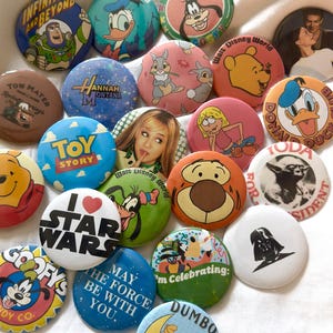 May include: A collection of colorful round buttons featuring various cartoon characters and movie themes. Designs include Buzz Lightyear, Donald Duck, Goofy, Winnie the Pooh, and Star Wars. Some buttons have text like "Toy Story" and "May the Force Be With You."