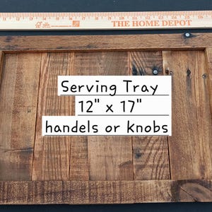 May include: A rectangular wooden serving tray with a natural brown finish, featuring vertical wood planks and two curved metal handles attached to the sides. The tray measures 12 inches by 17 inches, as indicated by the text and ruler.