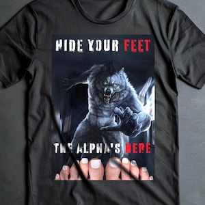 Puede incluir: Camiseta negra con un gráfico de un hombre lobo gruñendo y el texto "HIDE YOUR FEET THE ALPHA'S HERE". El diseño incluye un par de pies con uñas pintadas. La camiseta tiene mangas cortas.
