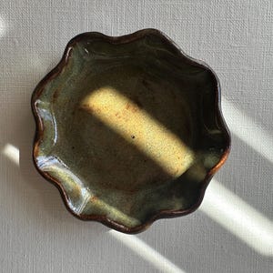 Pode incluir: Uma tigela de cerâmica feita à mão com uma borda ondulada única. A tigela apresenta uma mistura de tons verdes e castanhos, com uma superfície texturizada. O interior tem uma cor castanho-amarelada mais clara. Esta tigela decorativa é ideal para servir petiscos.