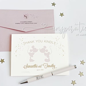 Puede incluir: Una tarjeta de agradecimiento rosa p&aacute;lido con siluetas de Mickey y Minnie Mouse, texto dorado y un sobre a juego. La tarjeta dice "Thank You Kindly" y est&aacute; personalizada con nombres y una fecha. Tambi&eacute;n hay un bol&iacute;grafo con "INSPIRE".