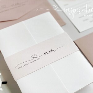 May include: Wedding invitation suite featuring a white card with a blush pink belly band. The band has a heart and initials, with the names and date below. Other blush pink envelopes and cards are visible.