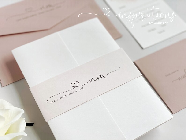 May include: Wedding invitation suite featuring a white card with a blush pink belly band. The band has a heart and initials, with the names and date below. Other blush pink envelopes and cards are visible.