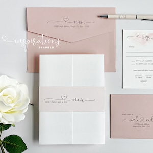 May include: A collection of wedding stationery in soft pink and white, including invitations, RSVP cards, and envelopes. The design features a heart motif and elegant script. A pen with the word "INSPIRE" is also included.