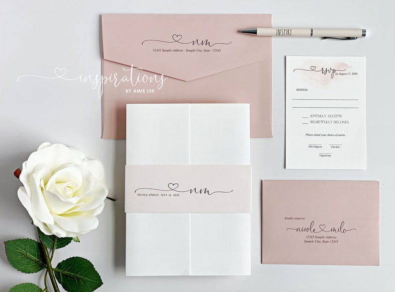 May include: A collection of wedding stationery in soft pink and white, including invitations, RSVP cards, and envelopes. The design features a heart motif and elegant script. A pen with the word "INSPIRE" is also included.