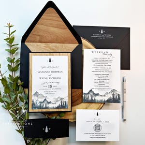 May include: Wedding invitation suite featuring a black and wood-toned envelope, invitation card, itinerary, and reply card. The invitation includes the names Susannah Hoffman and Wayne Richards, with a mountain landscape design. A pen is also included.