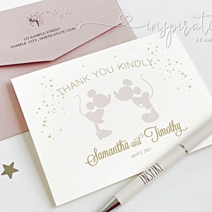 Puede incluir: Una tarjeta crema con letras doradas y un dise&ntilde;o de Mickey y Minnie Mouse, junto con un sobre rosa rubor. La tarjeta dice "Thank You Kindly" y "Samantha and Timothy, April 3, 2021." Tambi&eacute;n hay un bol&iacute;grafo con la palabra "INSPIRE".