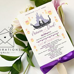 May include: A purple and white wedding program fan with a watercolor illustration of a couple in a gondola with lanterns and Cinderella's castle in the background. The text reads "Welcome to our fairy tale wedding" and "Mariessa and David".