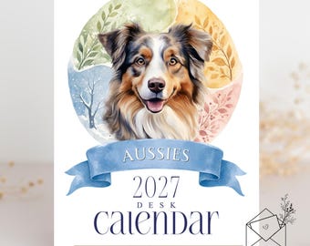 May include: A 2027 desk calendar featuring a watercolor illustration of an Australian Shepherd dog centered within a circular seasonal background. A blue ribbon banner displays the text AUSSIES above the year 2027 DESK CALENDAR.