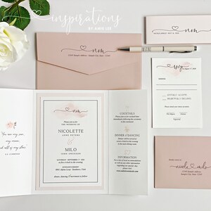 May include: A collection of blush pink wedding stationery, including invitations, RSVP cards, and envelopes. The design features a watercolor heart and the names "Nicole" and "Milo" in elegant script. A pen and a white rose are also present.