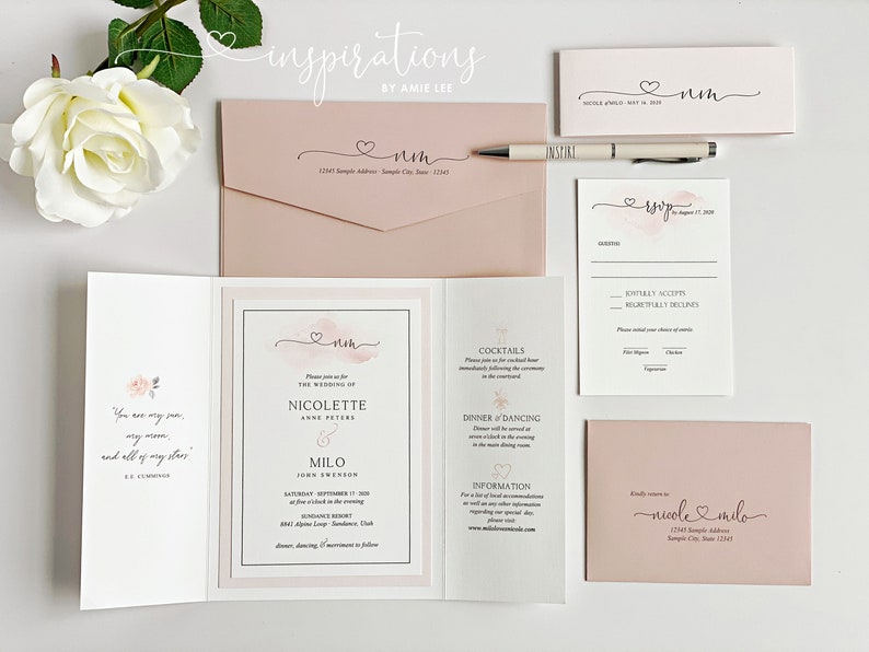 May include: A collection of blush pink wedding stationery, including invitations, RSVP cards, and envelopes. The design features a watercolor heart and the names "Nicole" and "Milo" in elegant script. A pen and a white rose are also present.