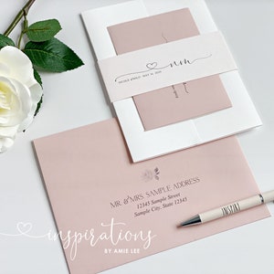 May include: Wedding invitation suite featuring a blush pink and white color scheme. The suite includes an invitation, envelope, and belly band with the couple's initials and wedding date. A pen with the word "INSPIRE" is also visible.