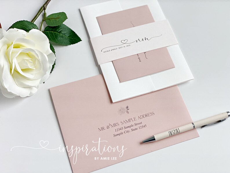 May include: Wedding invitation suite featuring a blush pink and white color scheme. The suite includes an invitation, envelope, and belly band with the couple's initials and wedding date. A pen with the word "INSPIRE" is also visible.