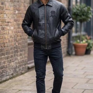 Puede incluir: Una chaqueta bomber de cuero negro con cremallera frontal, cuello en punta y pu&ntilde;os y dobladillo acanalados. La chaqueta presenta un emblema tonal sutil en el pecho izquierdo, combinada con pantalones vaqueros oscuros y zapatillas blancas.