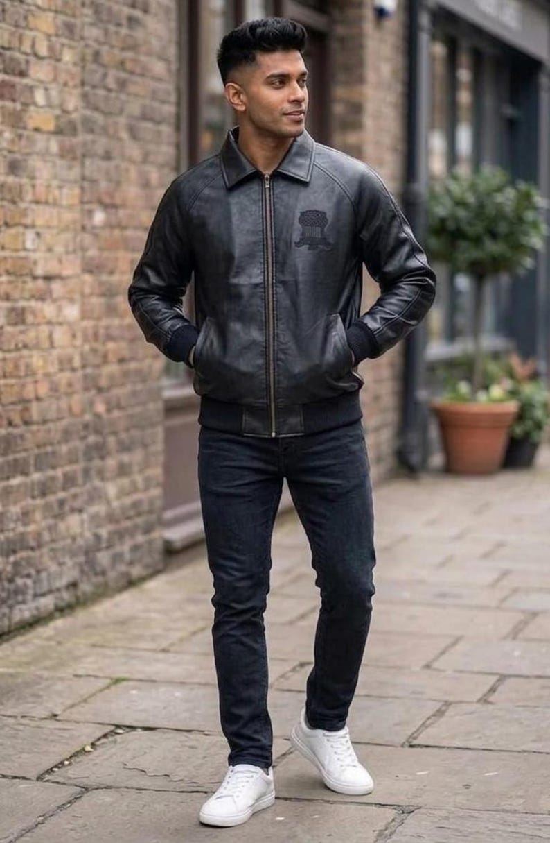 Puede incluir: Una chaqueta bomber de cuero negro con cremallera frontal, cuello en punta y pu&ntilde;os y dobladillo acanalados. La chaqueta presenta un emblema tonal sutil en el pecho izquierdo, combinada con pantalones vaqueros oscuros y zapatillas blancas.