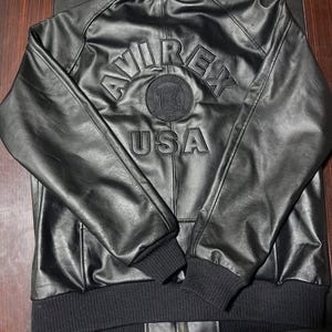 Puede incluir: Una chaqueta de cuero negra con cuello, pu&ntilde;os y dobladillo acanalados. La parte posterior muestra letras en relieve que dicen AVIREX y USA, con un emblema circular entre las palabras. La chaqueta tiene mangas largas y una cremallera frontal.