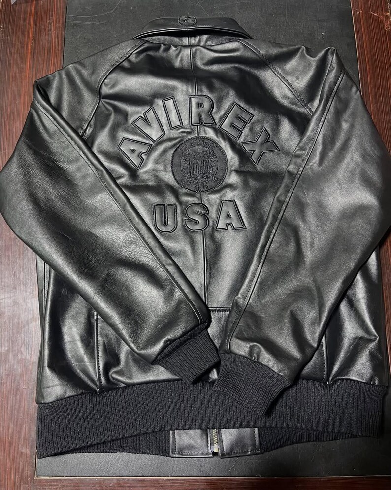 Puede incluir: Una chaqueta de cuero negra con cuello, pu&ntilde;os y dobladillo acanalados. La parte posterior muestra letras en relieve que dicen AVIREX y USA, con un emblema circular entre las palabras. La chaqueta tiene mangas largas y una cremallera frontal.