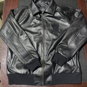 Puede incluir: Una chaqueta de cuero negra con cremallera frontal completa, cuello cl&aacute;sico y pu&ntilde;os y dobladillo de punto acanalado negro. El pecho muestra un parche bordado con el logotipo de Avirex en negro. La chaqueta tiene mangas largas.