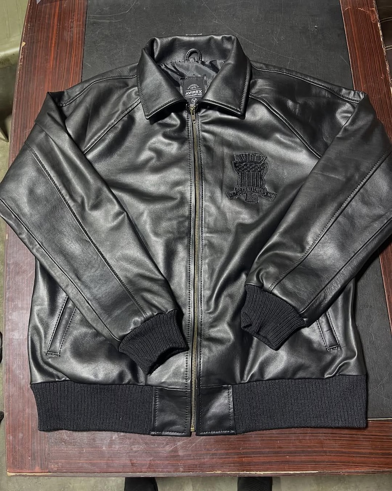 Puede incluir: Una chaqueta de cuero negra con cremallera frontal completa, cuello cl&aacute;sico y pu&ntilde;os y dobladillo de punto acanalado negro. El pecho muestra un parche bordado con el logotipo de Avirex en negro. La chaqueta tiene mangas largas.