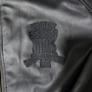 Puede incluir: Una chaqueta negra con un parche bordado en tono sobre tono en el pecho. El parche muestra el texto AVIREX en la parte superior, un dise&ntilde;o de escudo con estrellas y rayas, y una banda debajo que dice ESTABLISHED 1975 con NY a los lados.
