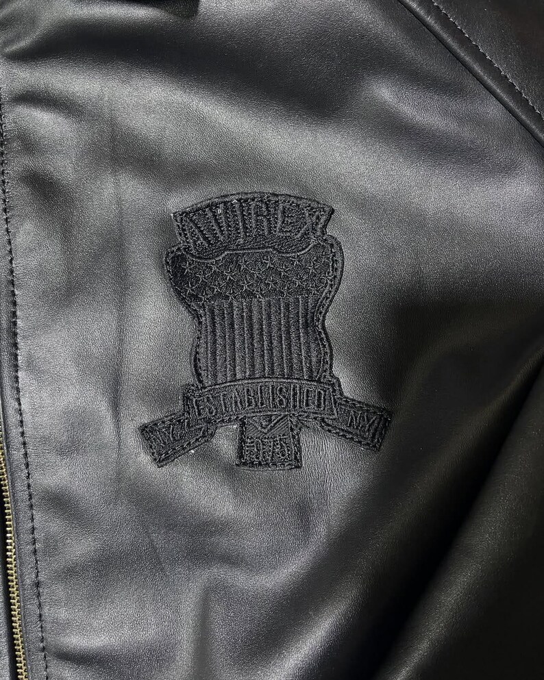 Puede incluir: Una chaqueta negra con un parche bordado en tono sobre tono en el pecho. El parche muestra el texto AVIREX en la parte superior, un dise&ntilde;o de escudo con estrellas y rayas, y una banda debajo que dice ESTABLISHED 1975 con NY a los lados.