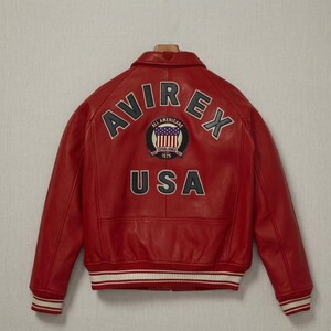 Op de afbeelding: Een rood leren bomberjack met een kraag, lange mouwen en geribbelde boorden en zoom met witte strepen. De achterkant toont de tekst AVIREX in een boog, een ronde patch met een vlagafbeelding en de tekst ALL AMERICANS ESTABLISHED 1975, en de tekst USA.