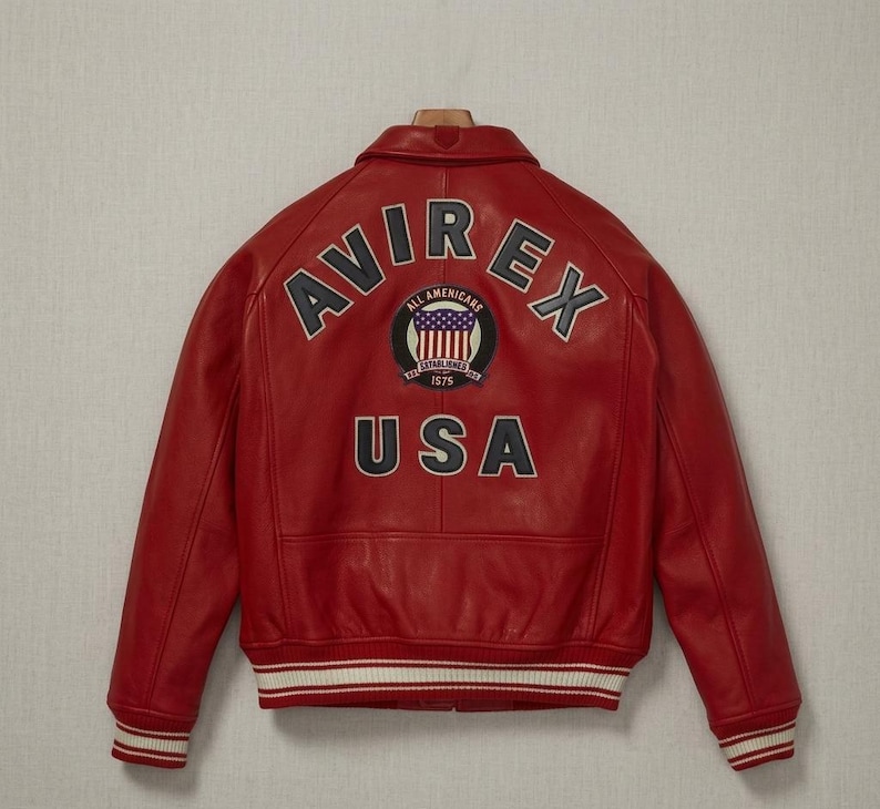 Op de afbeelding: Een rood leren bomberjack met een kraag, lange mouwen en geribbelde boorden en zoom met witte strepen. De achterkant toont de tekst AVIREX in een boog, een ronde patch met een vlagafbeelding en de tekst ALL AMERICANS ESTABLISHED 1975, en de tekst USA.
