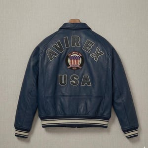 Op de afbeelding: Een marineblauwe leren bomberjack met de tekst AVIREX in een boog boven een ronde patch met een vlagontwerp en de tekst ALL AMERICANS ESTABLISHED 1975. Het woord USA staat onder de patch en het jack heeft gestreepte geribbelde boorden.