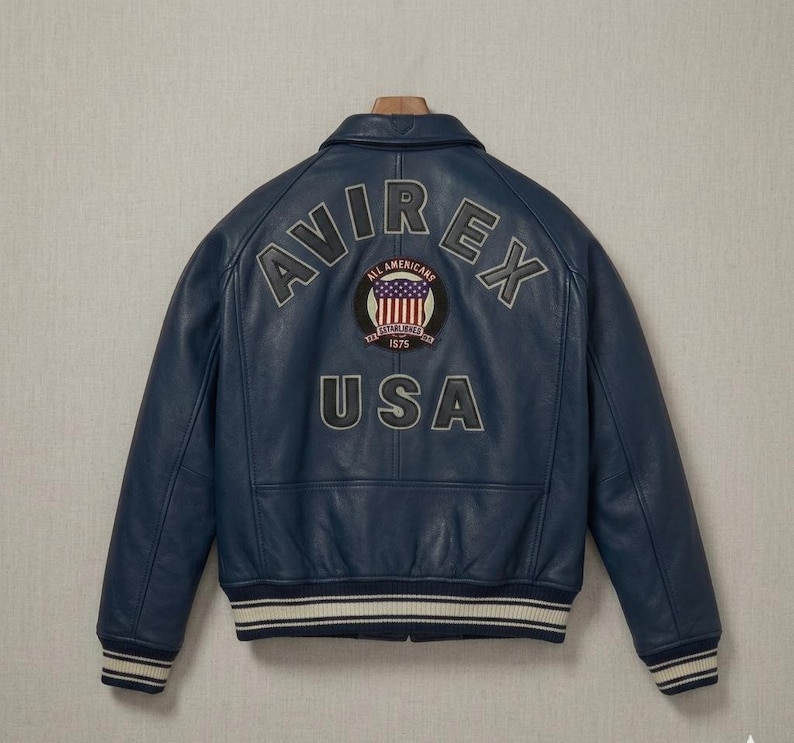 Op de afbeelding: Een marineblauwe leren bomberjack met de tekst AVIREX in een boog boven een ronde patch met een vlagontwerp en de tekst ALL AMERICANS ESTABLISHED 1975. Het woord USA staat onder de patch en het jack heeft gestreepte geribbelde boorden.