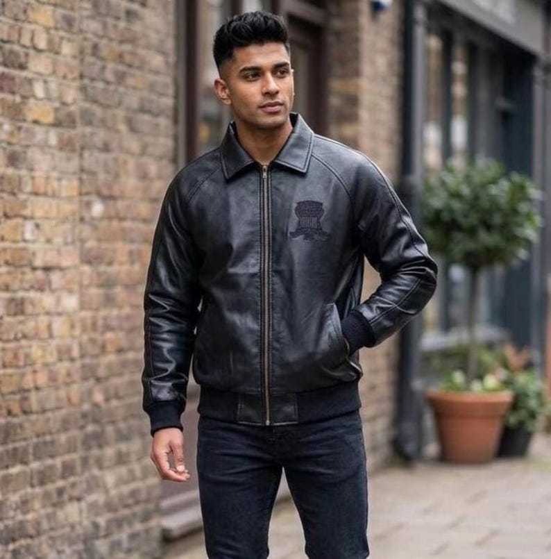 Puede incluir: Una chaqueta bomber de cuero negro con cuello cl&aacute;sico, cremallera frontal completa y pu&ntilde;os acanalados. La prenda presenta un emblema bordado oscuro y sutil en el lado izquierdo del pecho, combinada con pantalones vaqueros de mezclilla oscura.